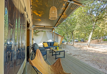 Udendørs opholdsområde foran et safaritelt med komfortable møbler i en skyggefuld skovcampingplads.