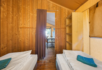 Vista interior de una tienda safari con dos camas individuales, estantería y cortina hacia comedor.