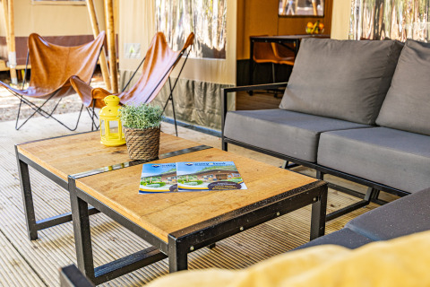 Gemütliche Outdoor-Lounge im Safari-Zelt mit Sesseln, Sofatisch, Deko-Pflanze und Prospekt auf dem Tisch.