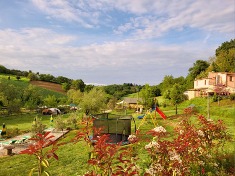 Panorama al Camping Suasa nelle Marche, Italia, con area giochi, trampolino e campagna verde sotto cielo azzurro.