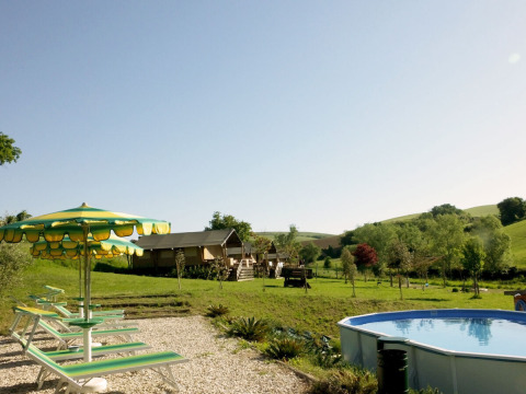 View of Camping Suasa in Marche, Italy, featuring a pool, loungers, umbrellas, and scenic green hills.