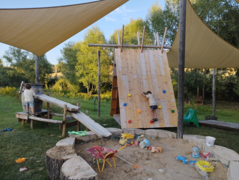 Kids play on a climbing wall and in a sandbox under sun shades at Camping Suasa holiday park in Italy.