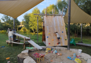 Kids play on a climbing wall and in a sandbox under sun shades at Camping Suasa holiday park in Italy.