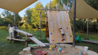 Kids play on a climbing wall and in a sandbox under sun shades at Camping Suasa holiday park in Italy.