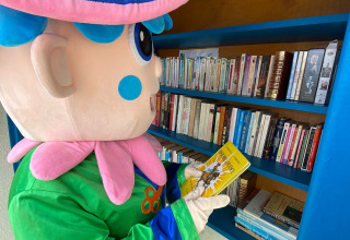 Mascotte in kleurrijk kostuum leest een boek bij een boekenplank op Flower Camping Le Pré des Sables, Frankrijk.
