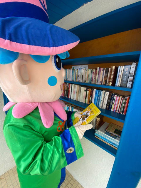 Mascotte in kleurrijk pak leest een boek aan een boekenrek bij Flower Camping Le Pré des Sables, Frankrijk.