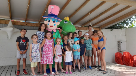 A group of children and adults pose with two mascots under a sheltered area at a holiday park in France.