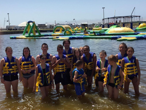 Un grupo de niños y adultos con chalecos salvavidas está de pie en el agua frente a un parque acuático inflable.