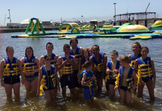 A group of children and adults in life jackets stand in the water near an inflatable obstacle course.