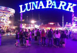 Letrero brillante de LUNAPARK en la entrada de un parque de atracciones de noche con gente reunida.