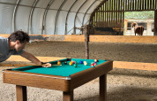 Man playing pool on a table inside a barn with a horse in the background at Feather Down Ecuries de Gapennes.