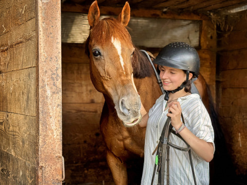 Een meisje met rijhelm naast een bruin paard in een houten stal bij Feather Down Ecuries de Gapennes in Frankrijk.