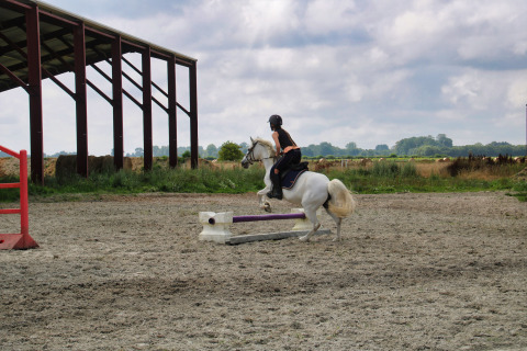 Rider jumps white horse over obstacle on outdoor arena at Feather Down Ecuries de Gapennes in France.