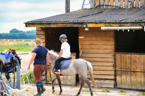 Child riding a pony guided by an adult in front of a wooden stable at Feather Down Ecuries de Gapennes.