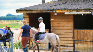 Kind rijdt op een pony onder begeleiding van een volwassene voor de stal bij Feather Down Ecuries de Gapennes.