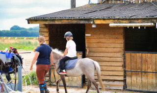 Kind rijdt op een pony onder begeleiding van een volwassene voor de stal bij Feather Down Ecuries de Gapennes.