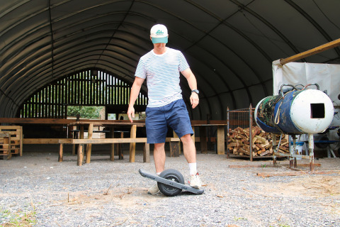 En mand med shorts og stribet trøje står oven på en elektrisk onewheel-skateboard i en lade hos Feather Down Ecuries de Gapennes.