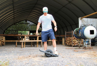 Een man met korte broek en gestreept shirt staat op een elektrische onewheel in een schuur bij Feather Down Ecuries de Gapennes.