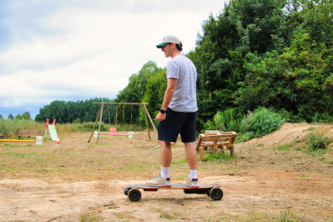 Mann auf Skateboard im Ferienpark mit Schaukeln, Rutsche und Sitzbank bei Feather Down Ecuries de Gapennes.