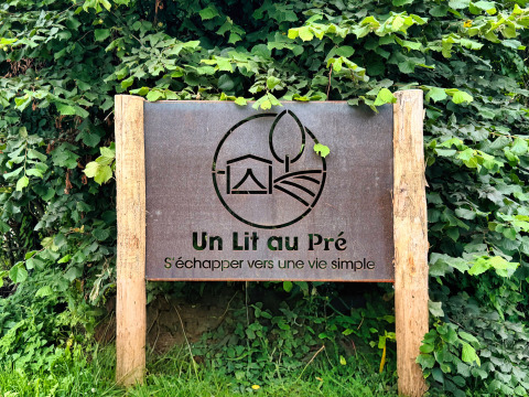 Cartello in legno con 'Un Lit au Pré' alle Ecuries de Gapennes, circondato da foglie verdi.