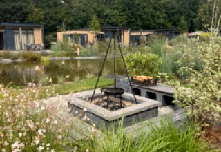 Fire pit and grill outside Batenburgh Deluxe lodge at Veluwepark de Bosgraaf, Netherlands, with cabins and greenery.