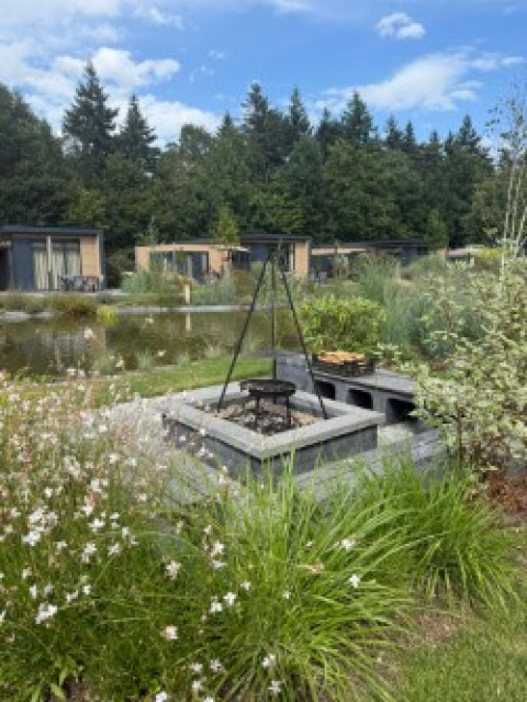 Feuerstelle und Grill vor den Batenburgh Deluxe Lodges im Veluwepark de Bosgraaf, Niederlande, mit Teich und Pflanzen.