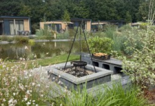 Feuerstelle und Grill vor den Batenburgh Deluxe Lodges im Veluwepark de Bosgraaf, Niederlande, mit Teich und Pflanzen.