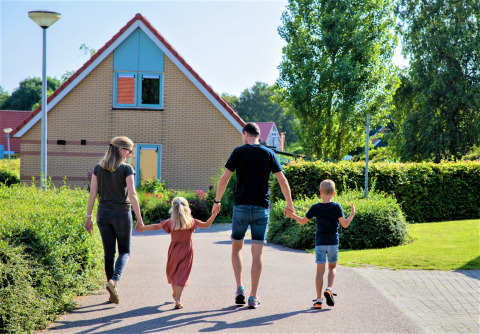 Une famille de quatre personnes marche main dans la main devant une maison entourée de verdure aux Pays-Bas.