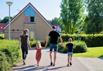 A family of four walks hand in hand along a path in front of a house surrounded by greenery in the Netherlands.