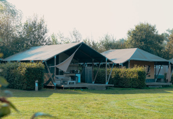 Safari cottage safari tent with shaded porch and greenery, set under clear skies on a bright day.