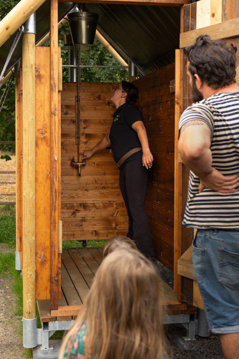 Femme montrant une douche extérieure à Feather Down Het Keelven, observée par un homme et un enfant.