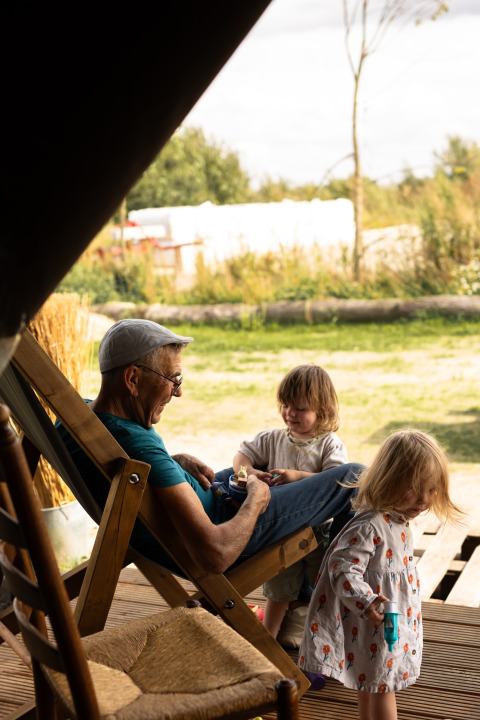 An elderly man relaxes in a deck chair outside while two small children play nearby at Feather Down Het Keelven.