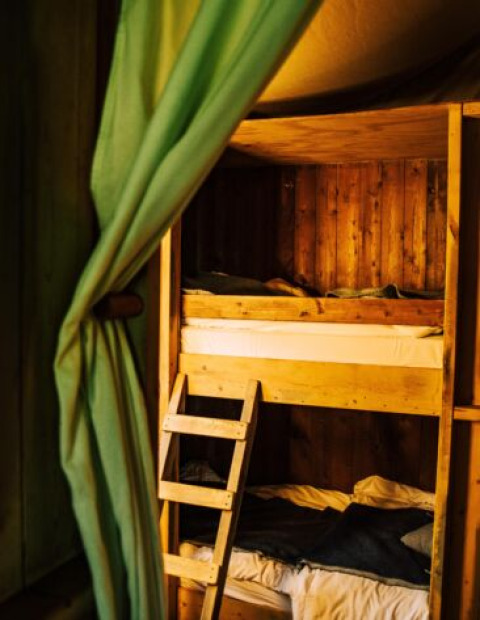 Intérieur de tente safari avec lit superposé en bois, rideau vert et lumière chaude et confortable.