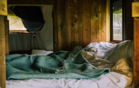 Gemütliches Bett mit Decke in einem rustikalen Safarizelt mit Holzverkleidung und Fenstern.
