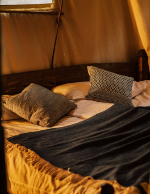 Letto accogliente con cuscini e coperta all'interno di una tenda safari, illuminata da luce calda.