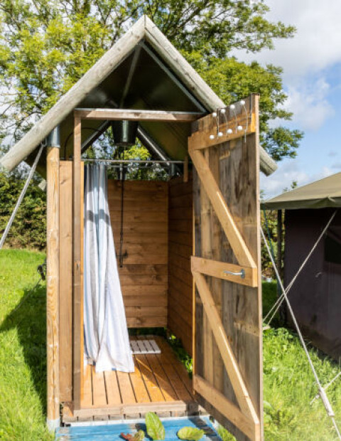 Douche extérieure à côté d'une tente safari, structure en bois avec porte ouverte sous ciel bleu et arbres.
