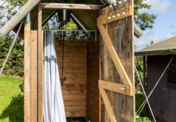 Doccia all'aperto accanto a una tenda safari, struttura in legno con porta aperta sotto cielo azzurro.