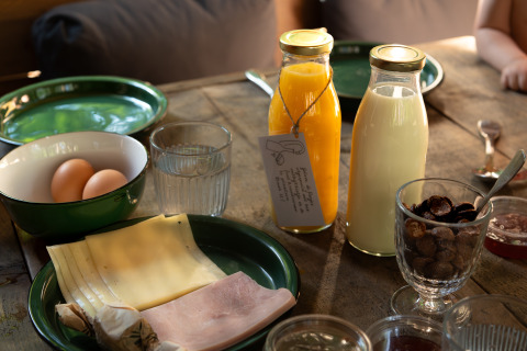 Glamping ontbijt op een houten tafel met sap, melk, eieren, kaas, ham en noten in kommetjes.