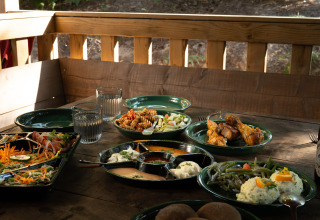 Tavolo all'aperto con insalate, pollo, bicchieri d'acqua e piatti vari presso una struttura glamping.