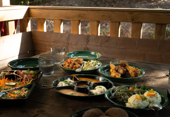 Buiten houten tafel met salades, kippenbouten, waterglazen en diverse gerechten bij een glamping verblijf.