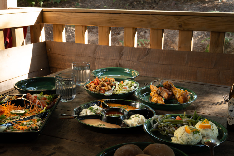 Outdoor wooden table with salads, chicken drumsticks, water glasses, and assorted dishes at a glamping site.