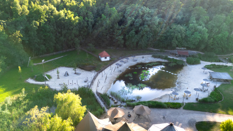 Vista aerea di un glamping con tende, ombrelloni, piccolo stagno, aree sabbiose e foresta sullo sfondo.