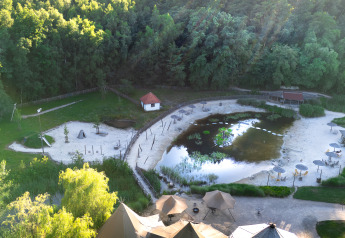 Luftaufnahme einer Glamping-Anlage mit Zelten, Sonnenschirmen, einem Teich und bewaldeter Umgebung.