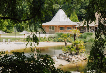 Hébergement glamping avec cabane en bois, palmiers, plage de sable et étang entouré de végétation luxuriante.