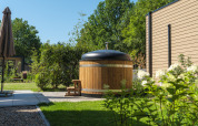 Outdoor wooden hot tub with black lid by a lodge, surrounded by a lush green garden and trees in sunlight.