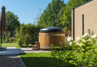 Outdoor wooden hot tub with black lid by a lodge, surrounded by a lush green garden and trees in sunlight.