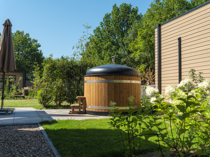 Buitenspa van hout met zwart deksel bij een lodge, omgeven door groene tuin en bomen op een zonnige dag.