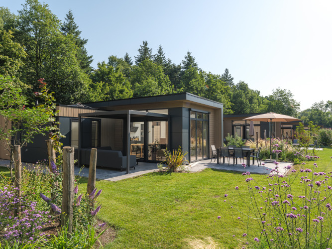 Moderna lodge Essenburgh Comfort con grandes ventanales, patio amueblado y jardín junto a un bosque.