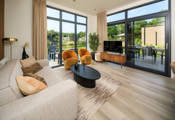 Moderne lodge-stue med store vinduer, beige sofa, gule stole og udsigt til have og terrasse om sommeren.