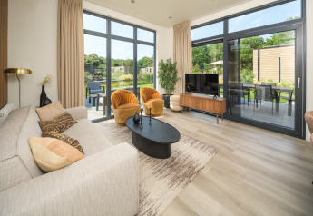 Modernes Lodge-Wohnzimmer mit großen Fenstern, gelbem Sessel und Blick auf Garten und Terrasse im Sommer.